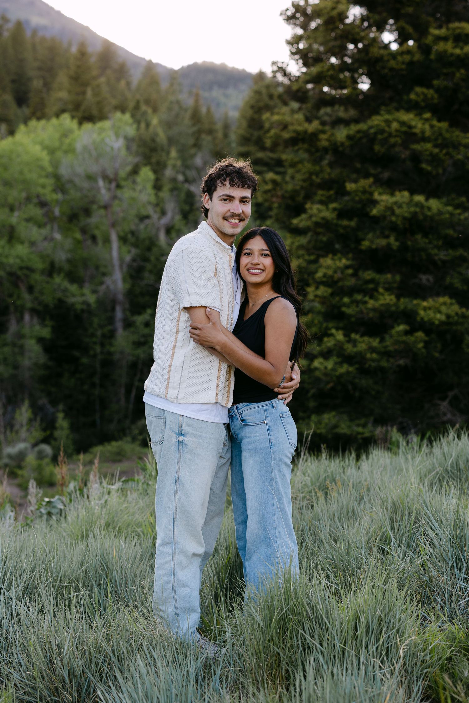 A smiling couple embraces in a lush green meadow with pine trees and mountains in the background.