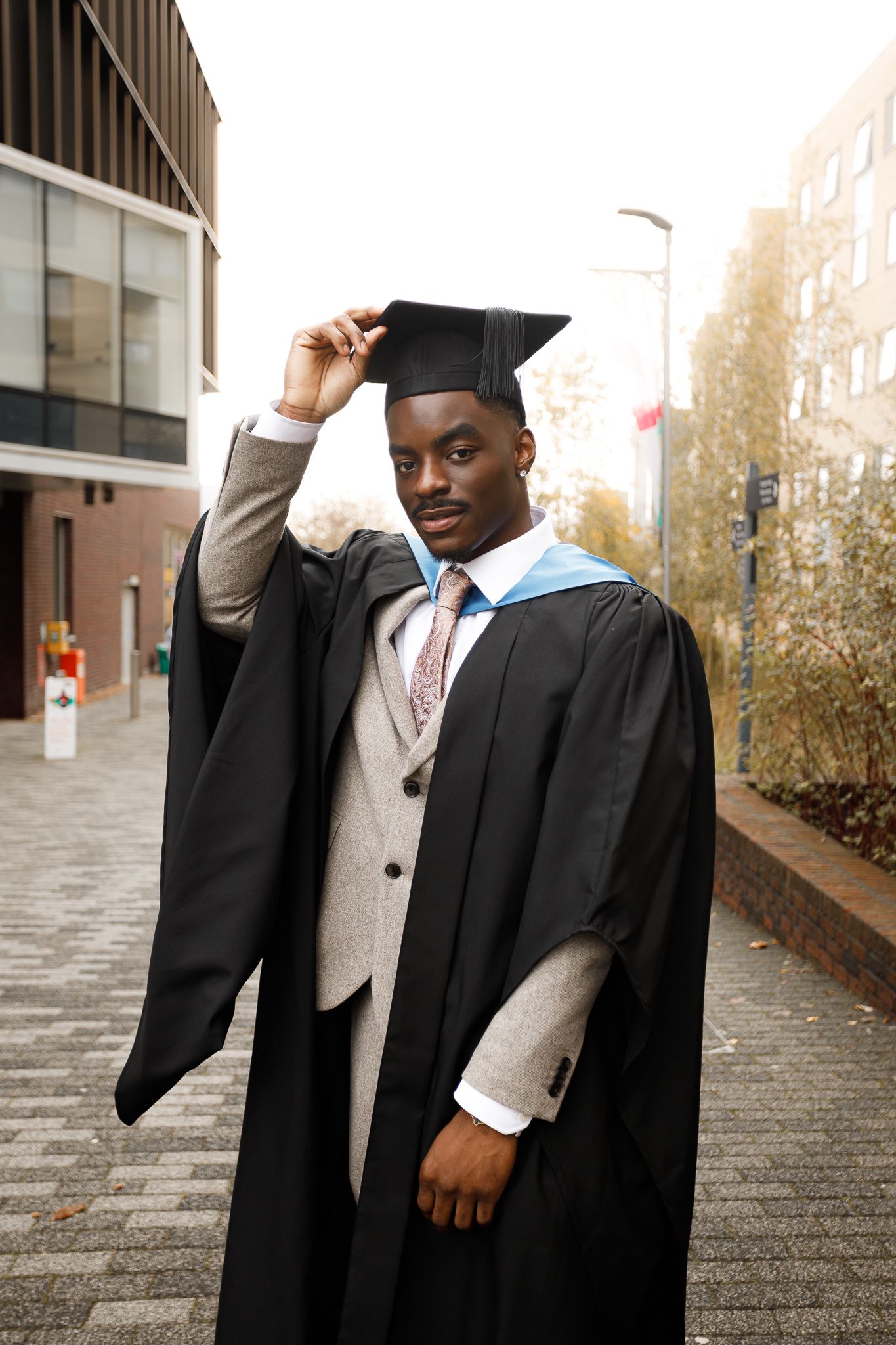 Graduate in academic regalia poses with graduation cap and gown on campus steps during commencement ceremony.
