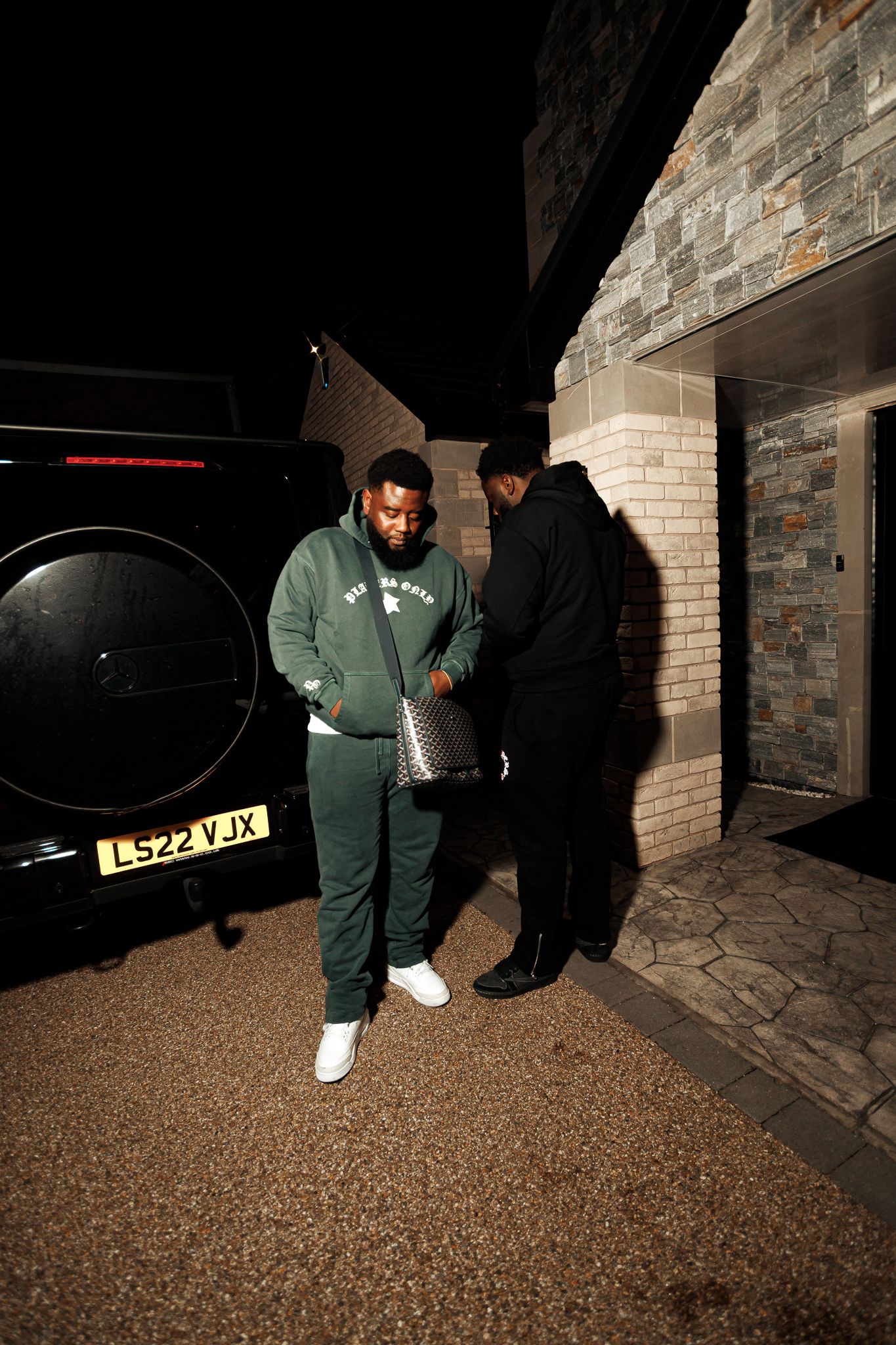 Two people stand outside a brick building at night next to a dark vehicle on a textured driveway.