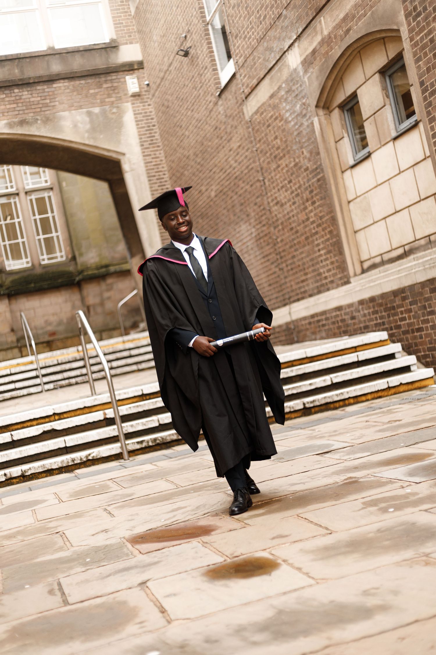Graduate in black cap and gown walks across stone courtyard during graduation ceremony at historic brick building.