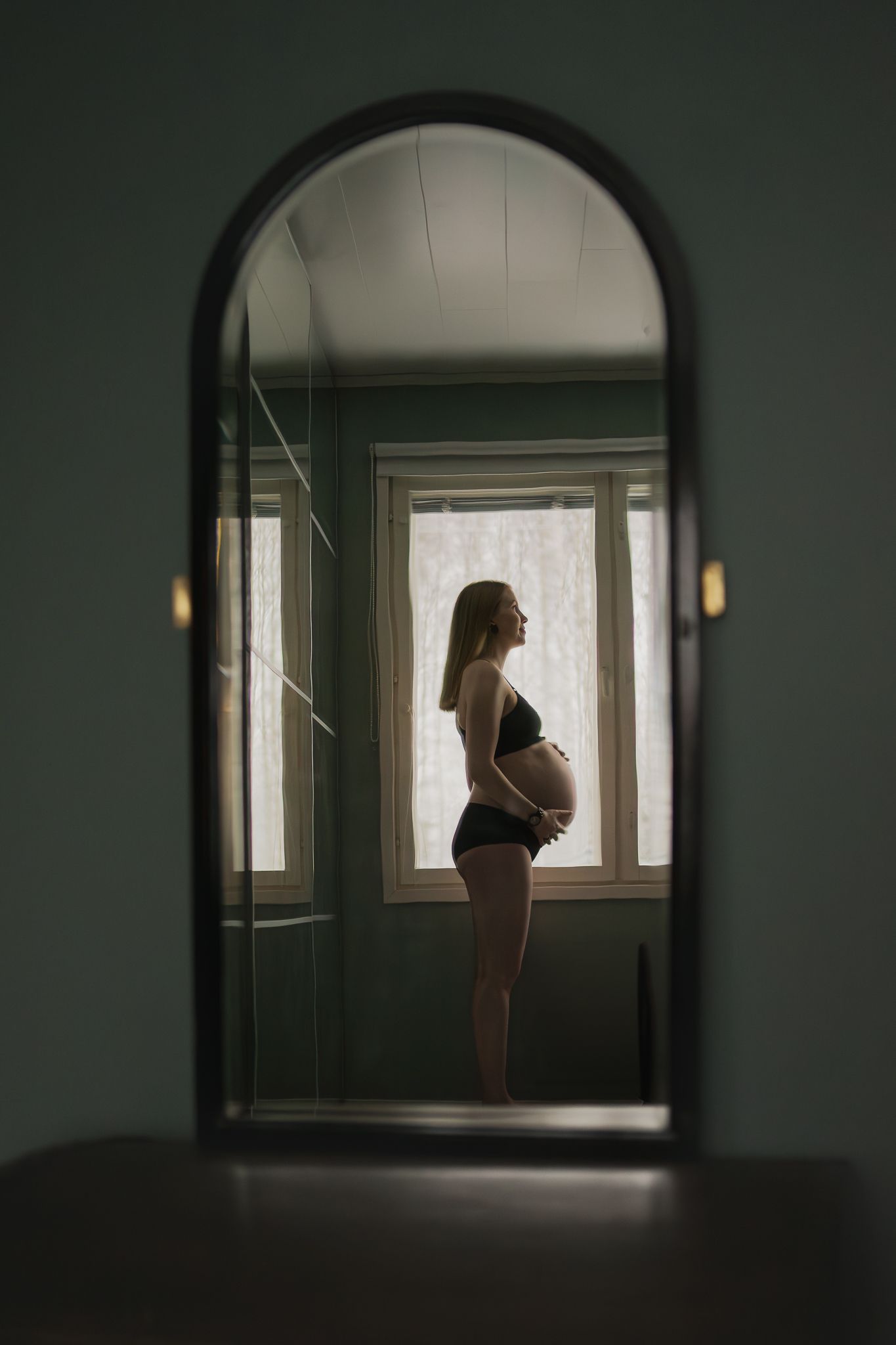 Silhouette of pregnant woman standing by window, viewed through arched doorway.