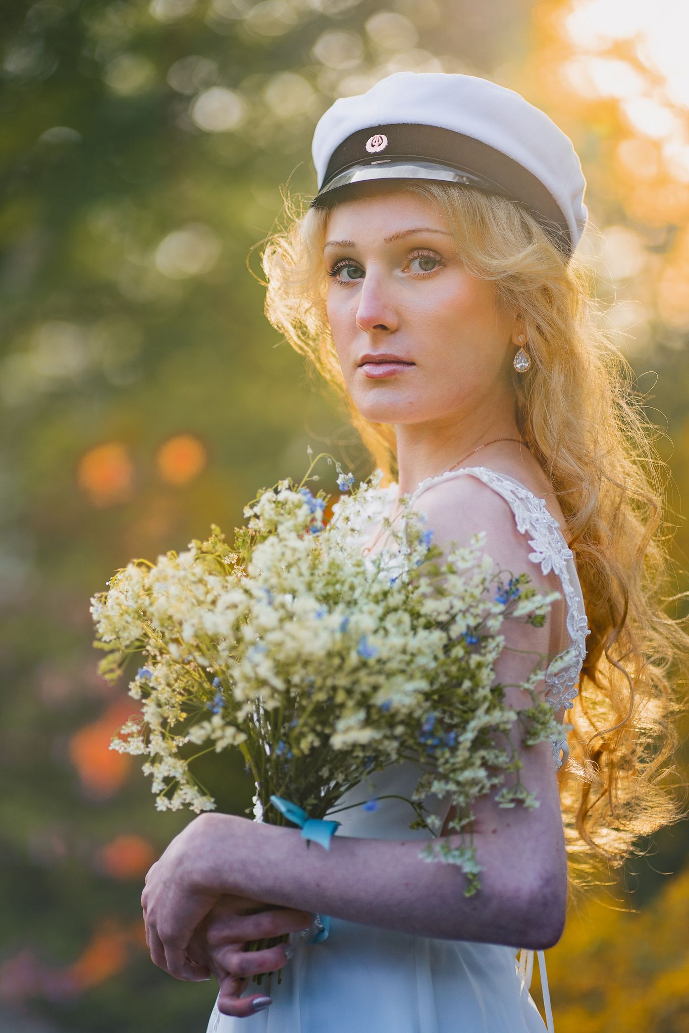 A dreamy portrait showing someone in a white dress and sailor cap holding a delicate bouquet at golden hour.