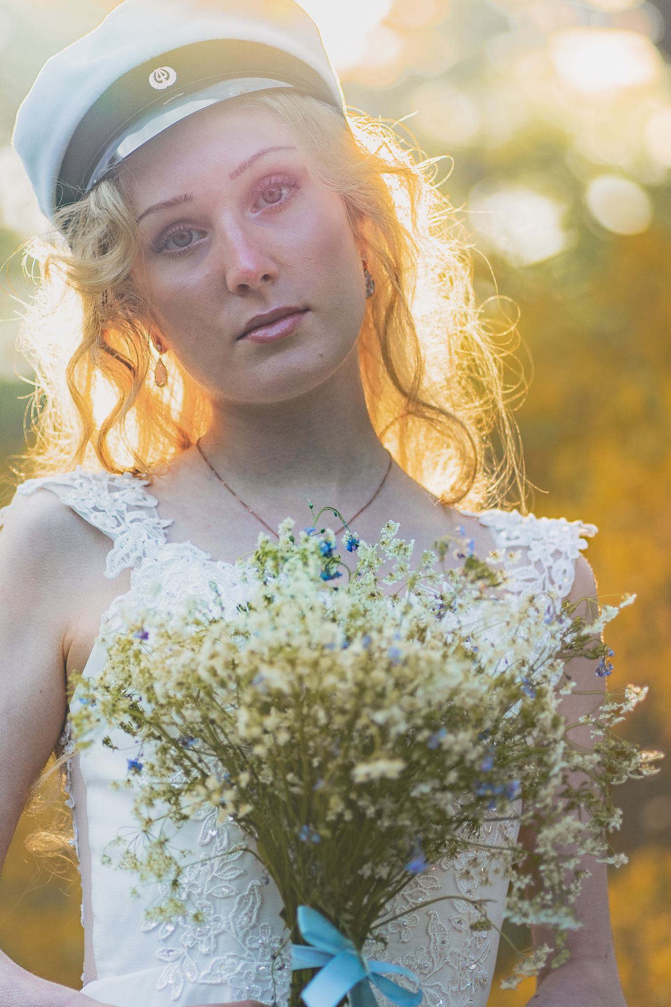 A bride in a white lace dress and nautical cap holds a delicate baby's breath bouquet with blue ribbon in golden sunset light.