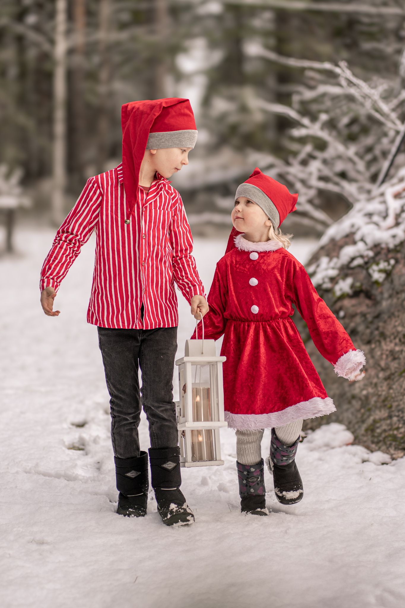 Two children in red winter outfits walking together on a snowy path while carrying a lantern.