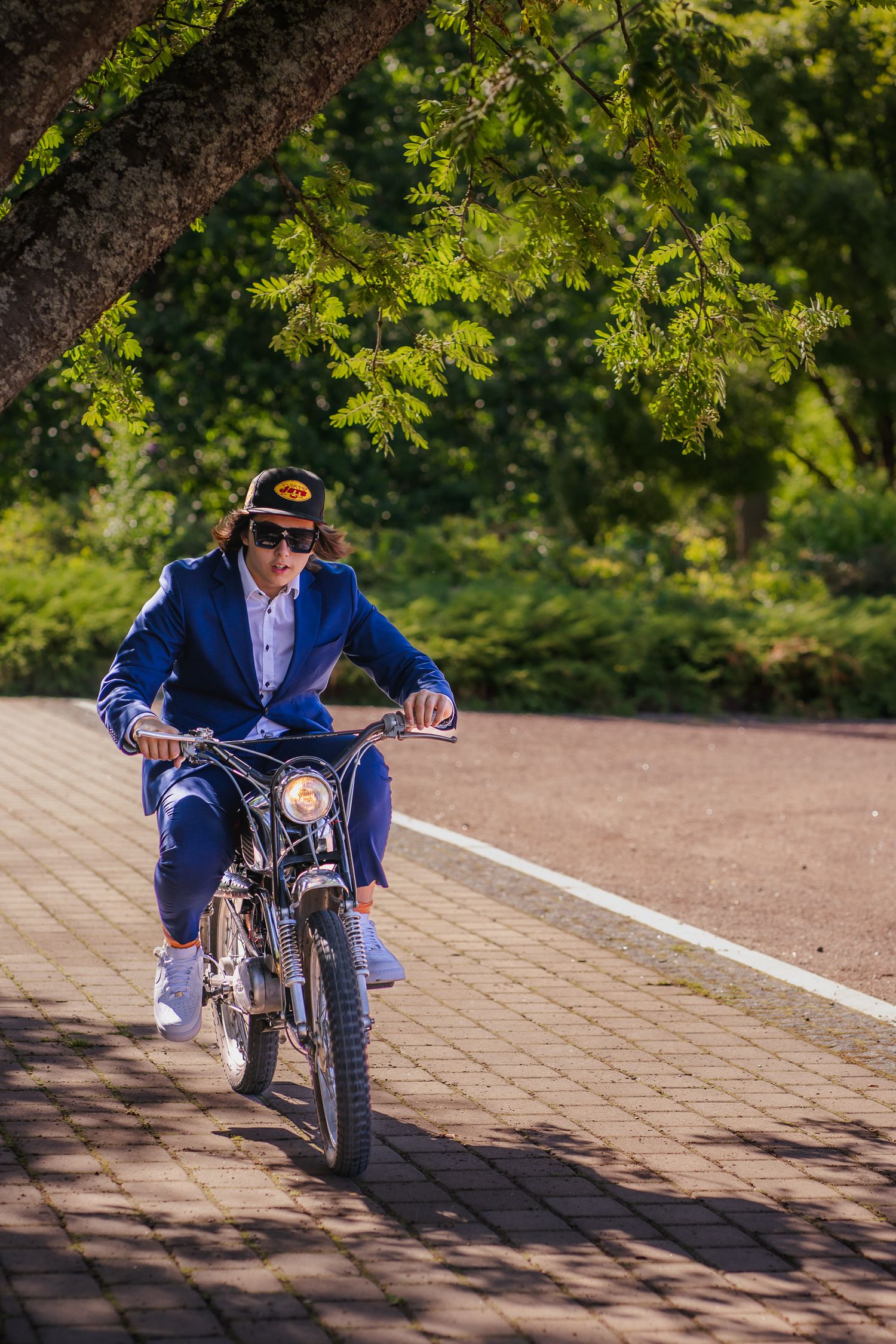 Person riding vintage motorcycle on brick path under green tree branches.