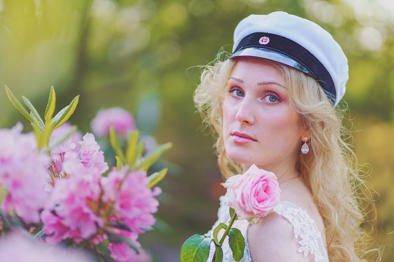 A person in a white graduation cap poses with pink roses and rhododendron flowers in a dreamy outdoor setting.