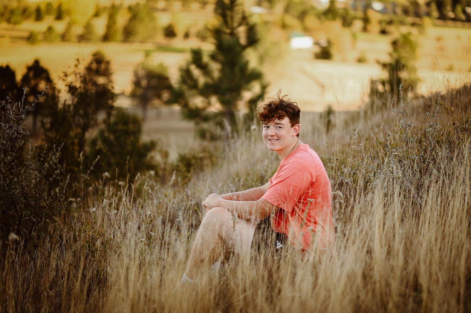 Someone in a coral colored shirt sits in golden dry grass during sunset with mountains visible in the background.