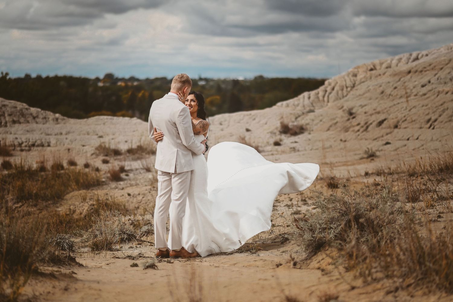 A couple in wedding attire embraces atop a sandy hill against moody cloudy skies as her white dress catches the wind.