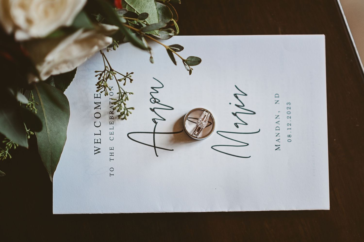 Wedding rings placed on a white invitation card with elegant calligraphy, surrounded by green foliage.