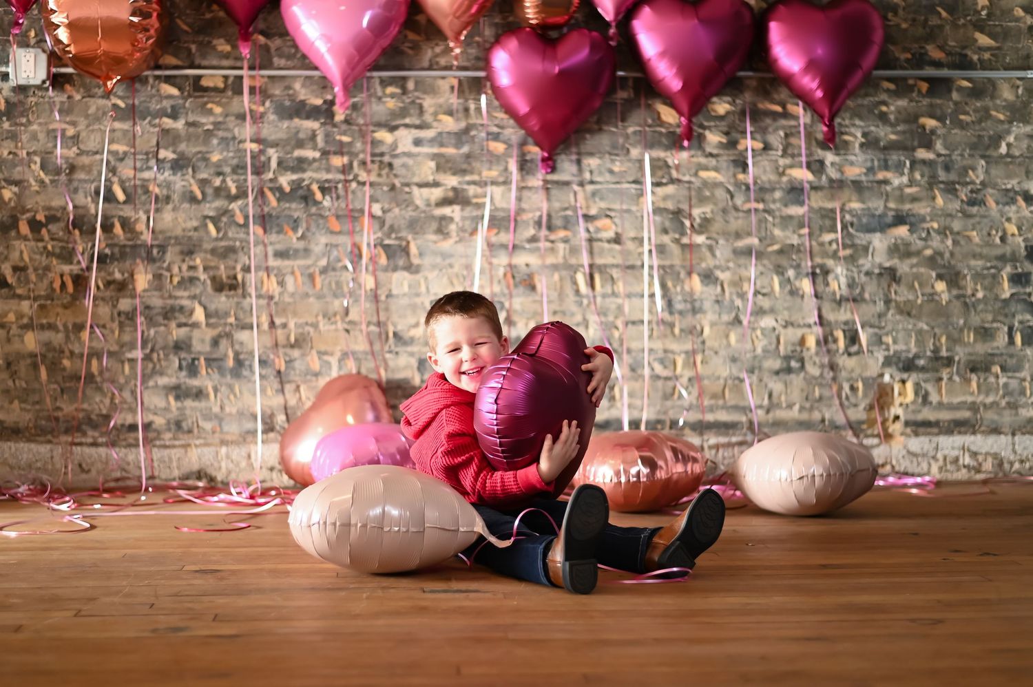 Pink and red heart balloons float against rustic brick wall with scattered props on wooden floor in Valentine's decor setup.