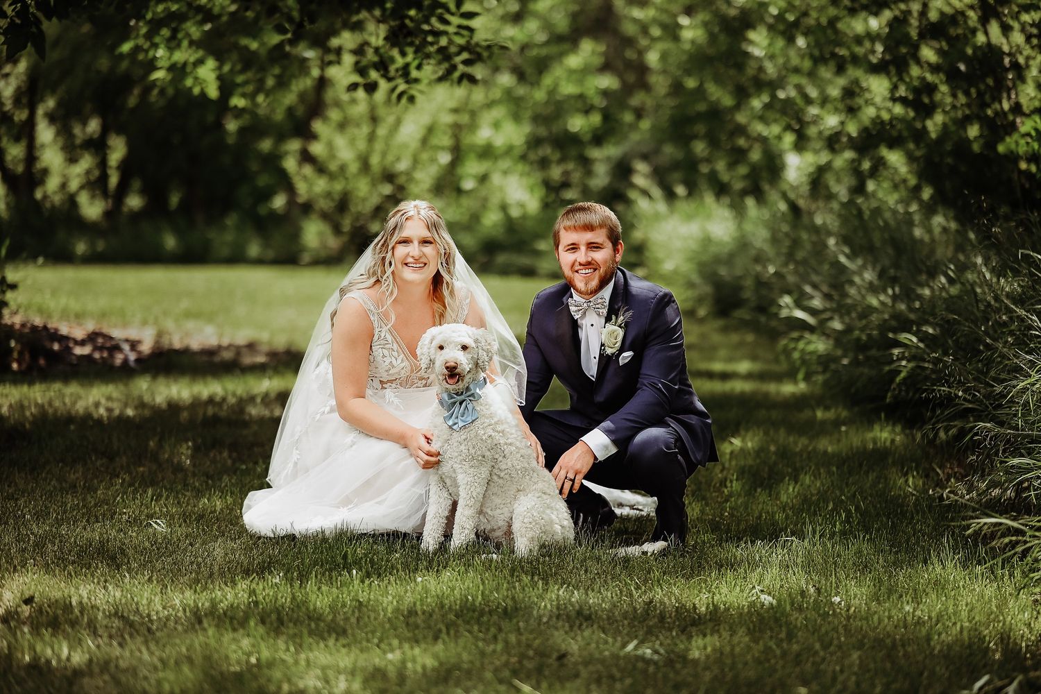 A newlywed couple poses with their fluffy white dog on a sunny lawn during their wedding photo session.