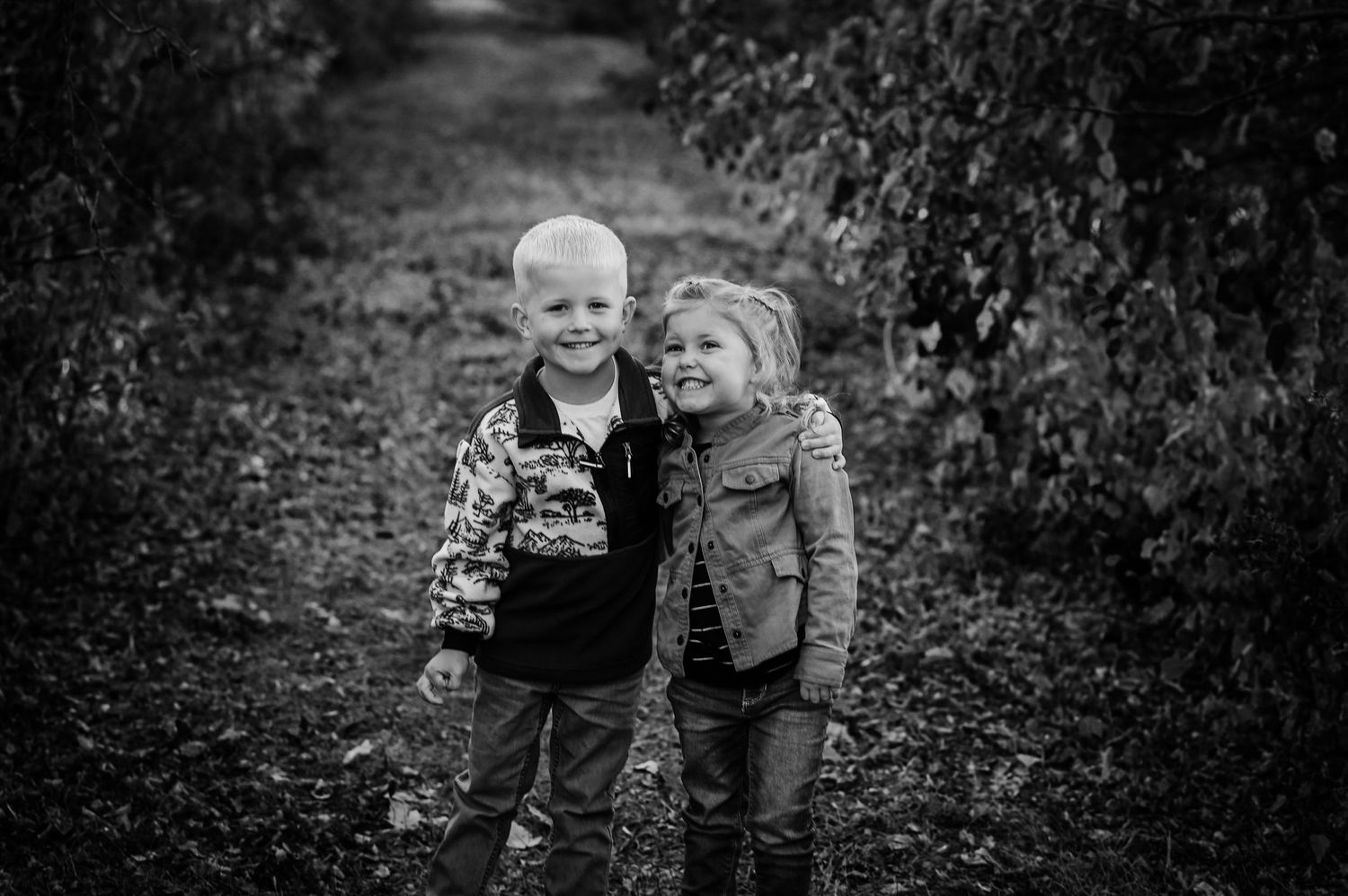 Two children stand together on a tree-lined path in black and white photography.