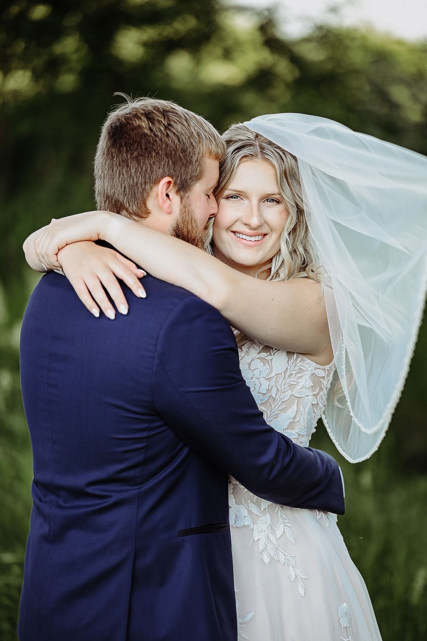 Newlywed couple embraces outdoors, bride wearing lace gown with veil and groom in navy suit.