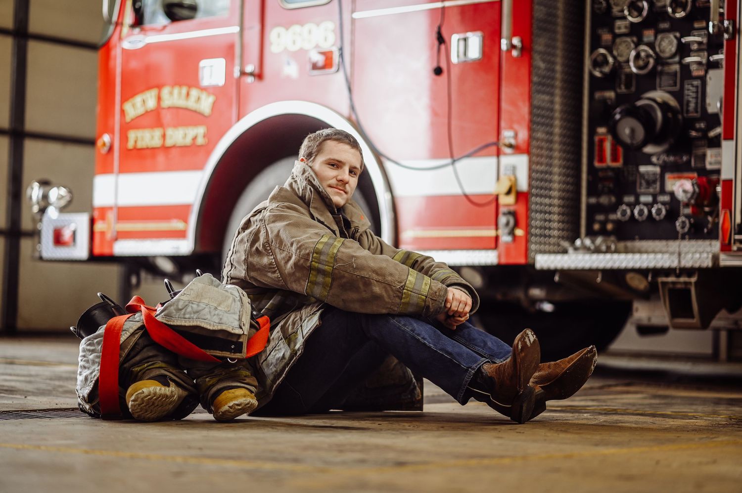 A firefighter rests on the ground next to their gear bag and red fire truck in a fire station.