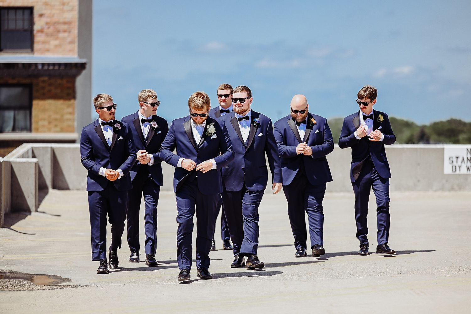 A group of groomsmen in matching navy suits and sunglasses walk together on a sunny day.