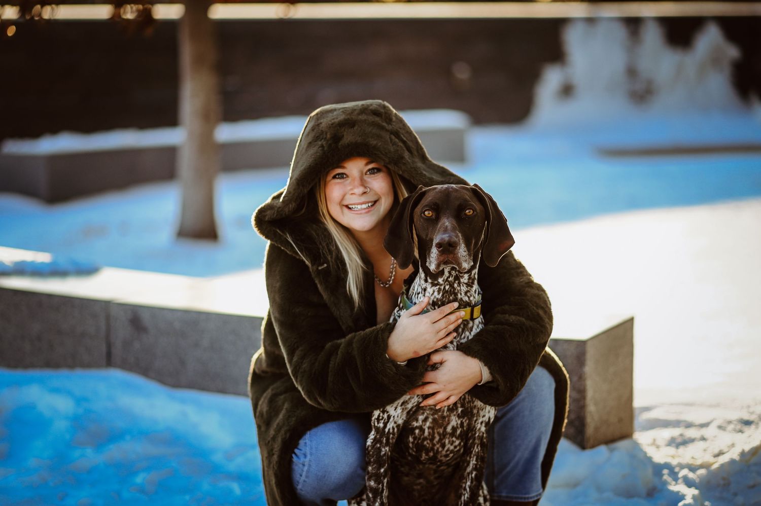 A person in a brown fur coat sits in the snow with their German Shorthaired Pointer dog on a winter day.