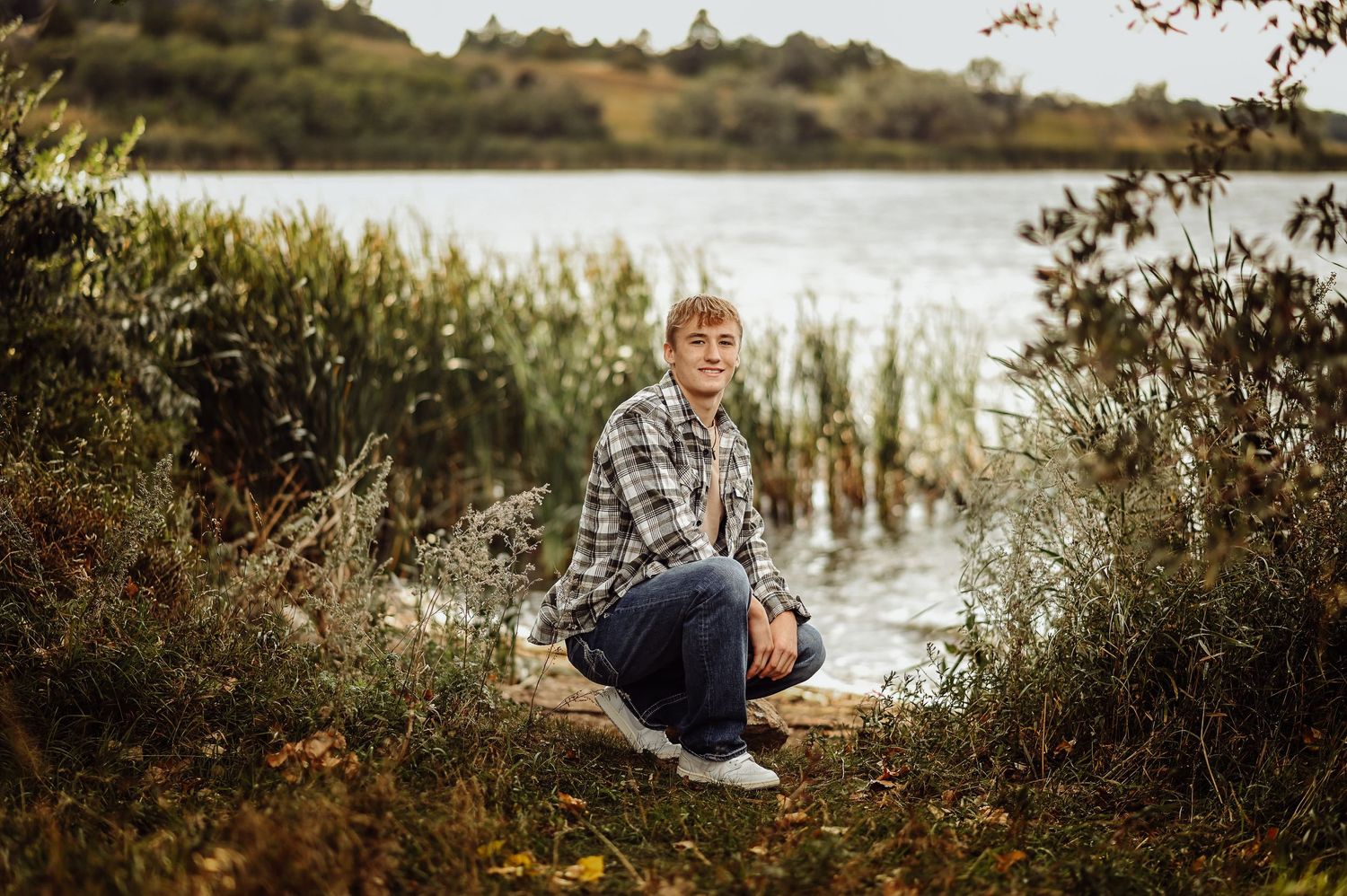 Senior boy in plaid sits on a fallen log by a tranquil lake surrounded by tall reeds at sunset.