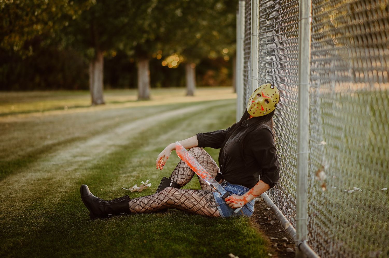 Halloween-themed portrait of someone in a yellow mask sitting against a chain-link fence at sunset.
