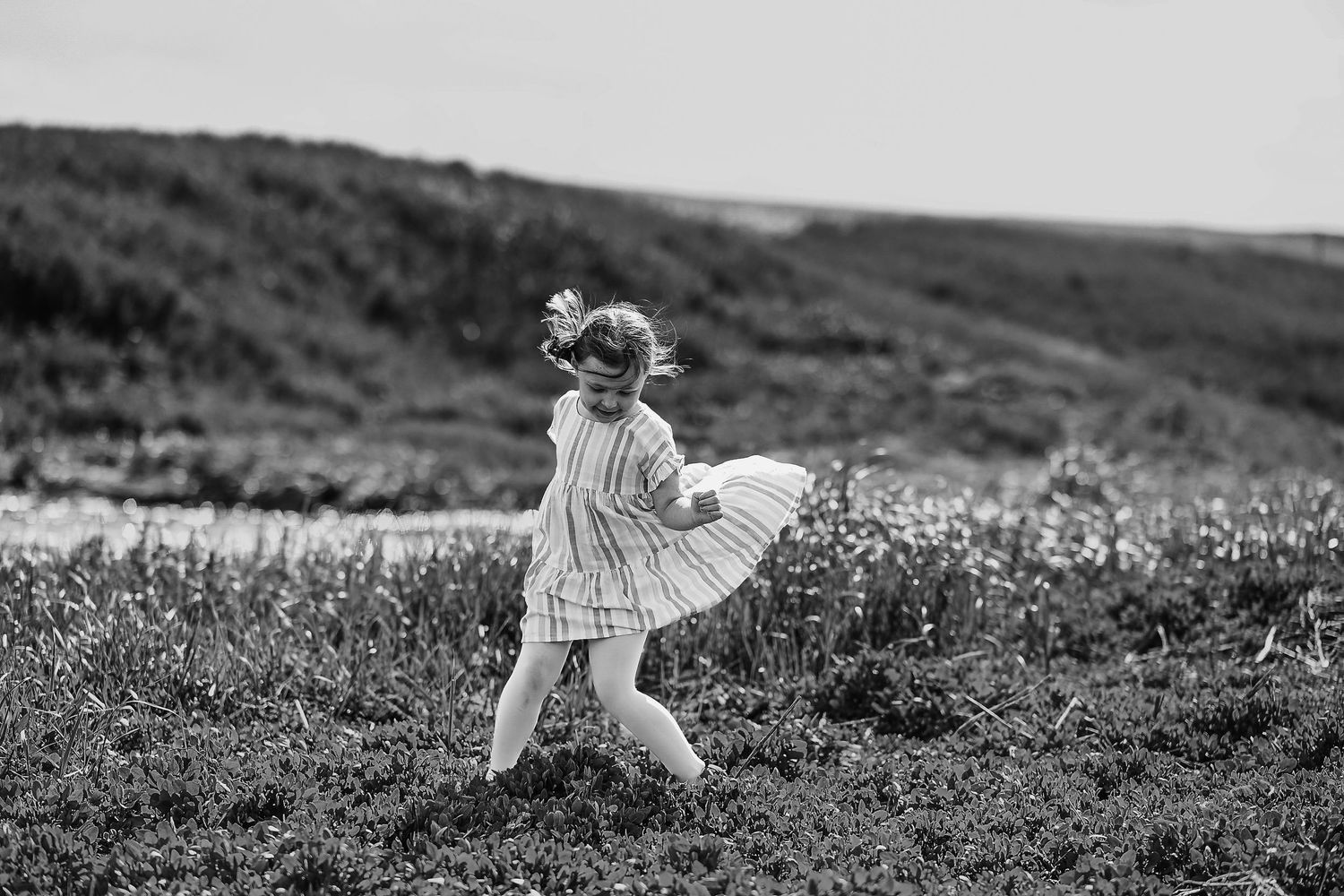 A child in a striped dress twirls joyfully in a meadow on a hillside in black and white.