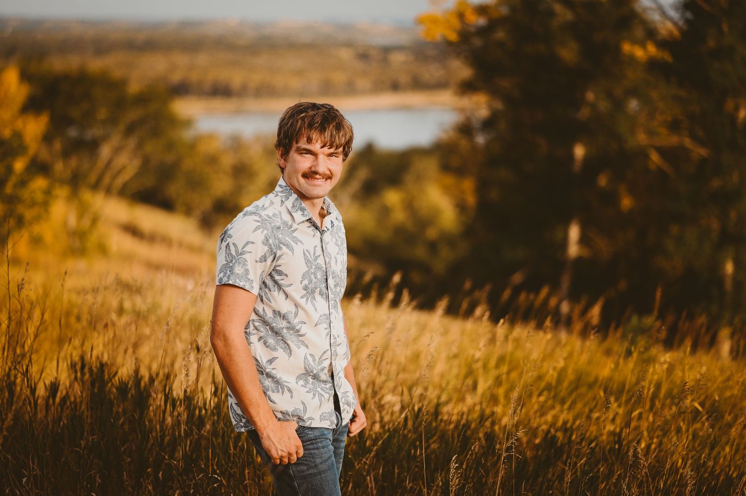 Senior boy in a floral print shirt stands in golden grass near a lake during sunset with autumn colors in background.