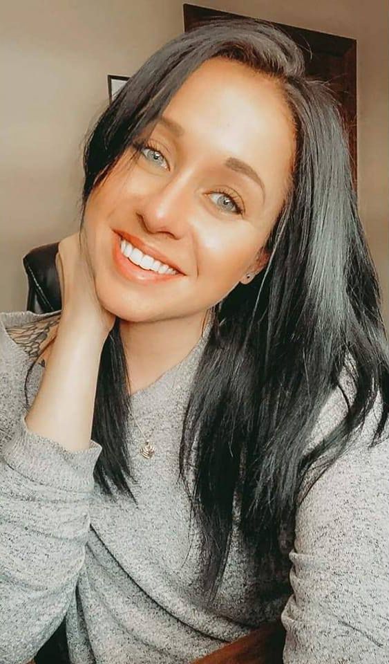 Selfie portrait showing bright smile and long dark hair wearing a casual grey sweater in warm lighting.