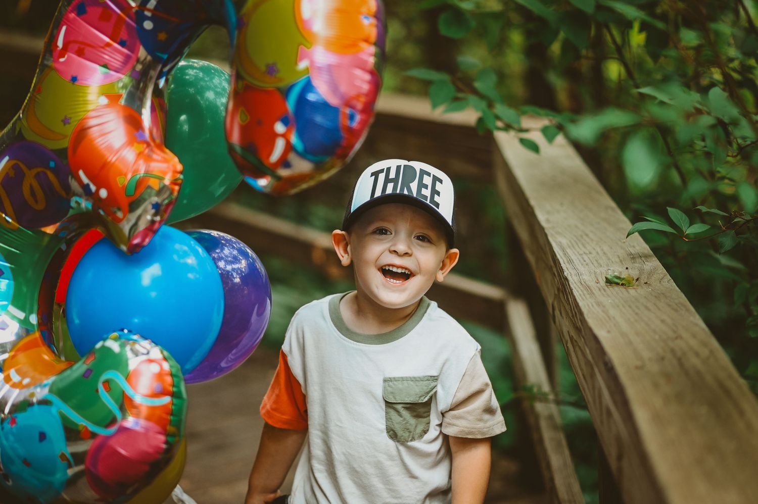 Colorful party balloons float beside a smiling child in a baseball cap on a wooden bench in an outdoor setting.