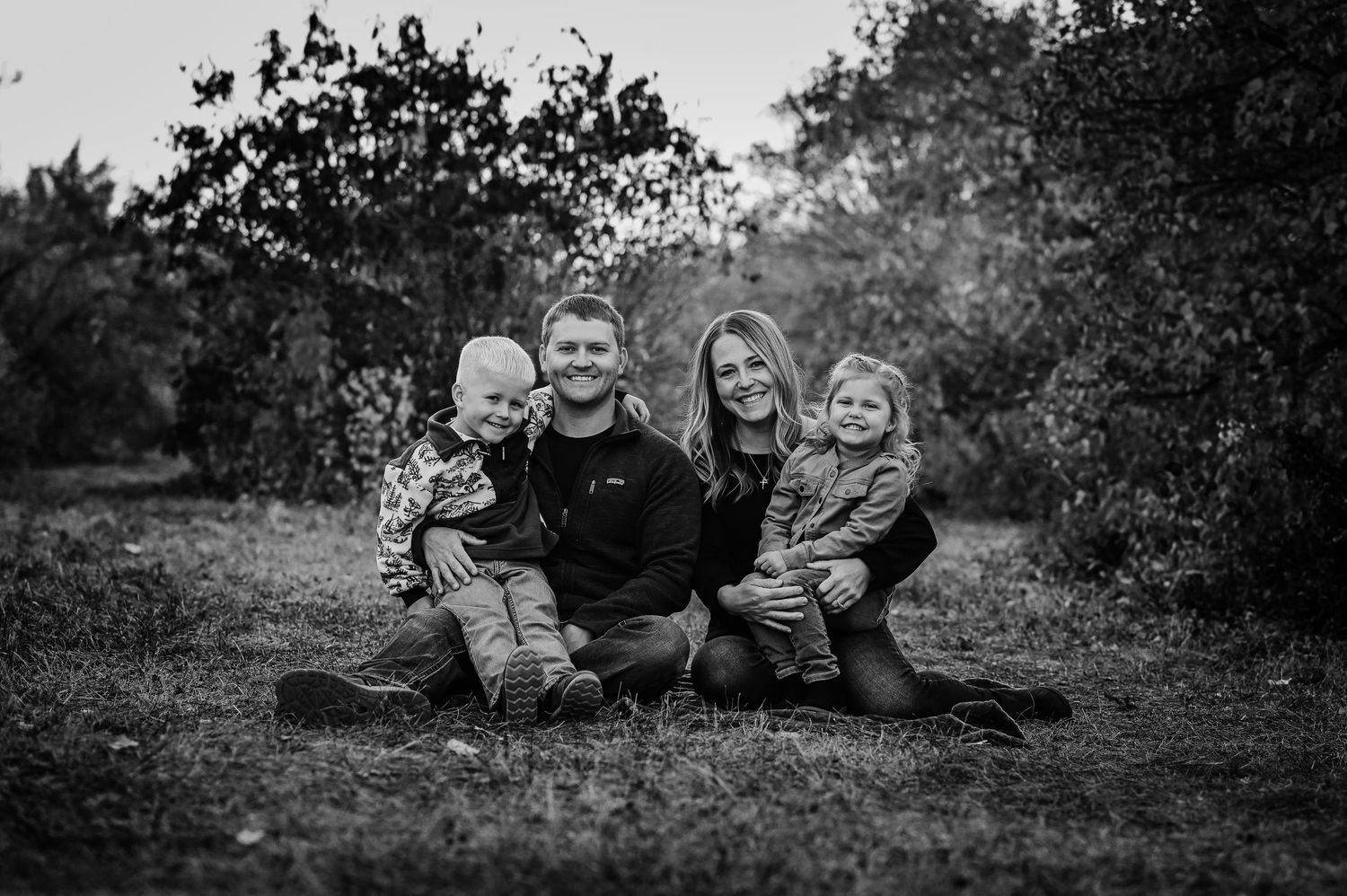 Black and white outdoor portrait of a family sitting together on the ground in a natural setting.