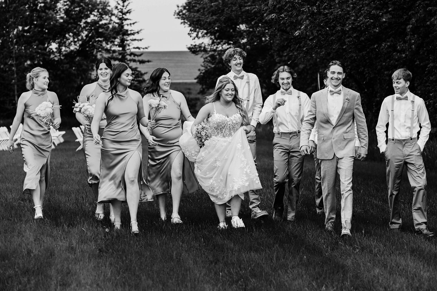 A wedding party runs joyfully through a grassy field in black and white, with bridesmaids in gray dresses and groomsmen in suspenders.