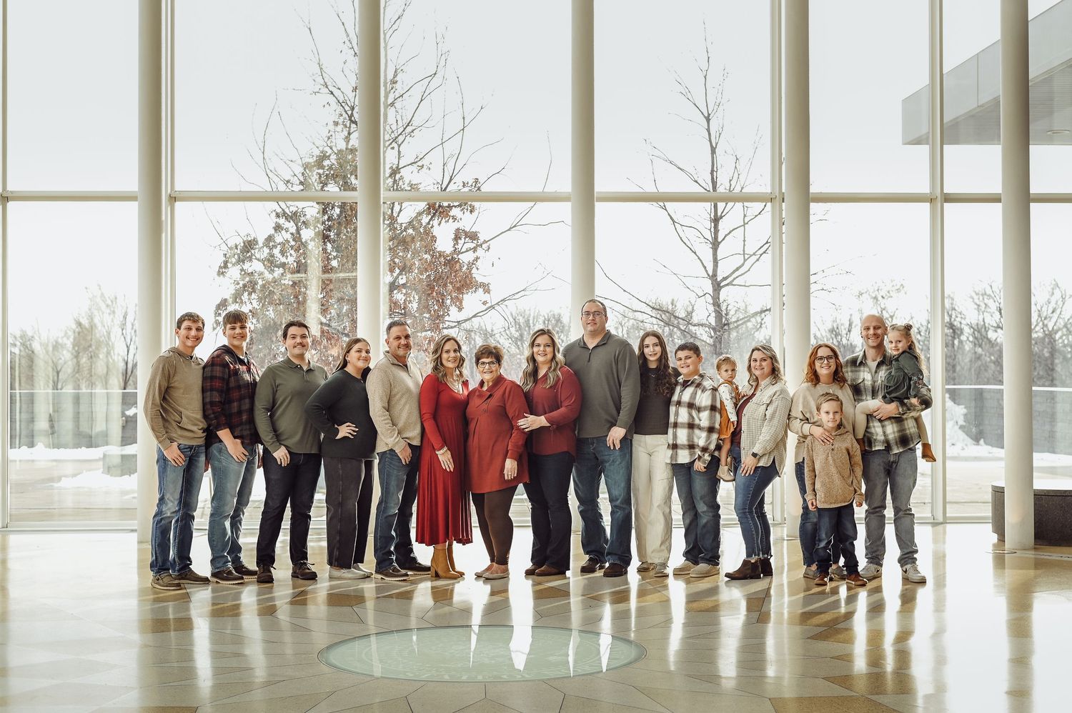 A large casually dressed family standing together in a bright sunlit room with floor to ceiling windows.