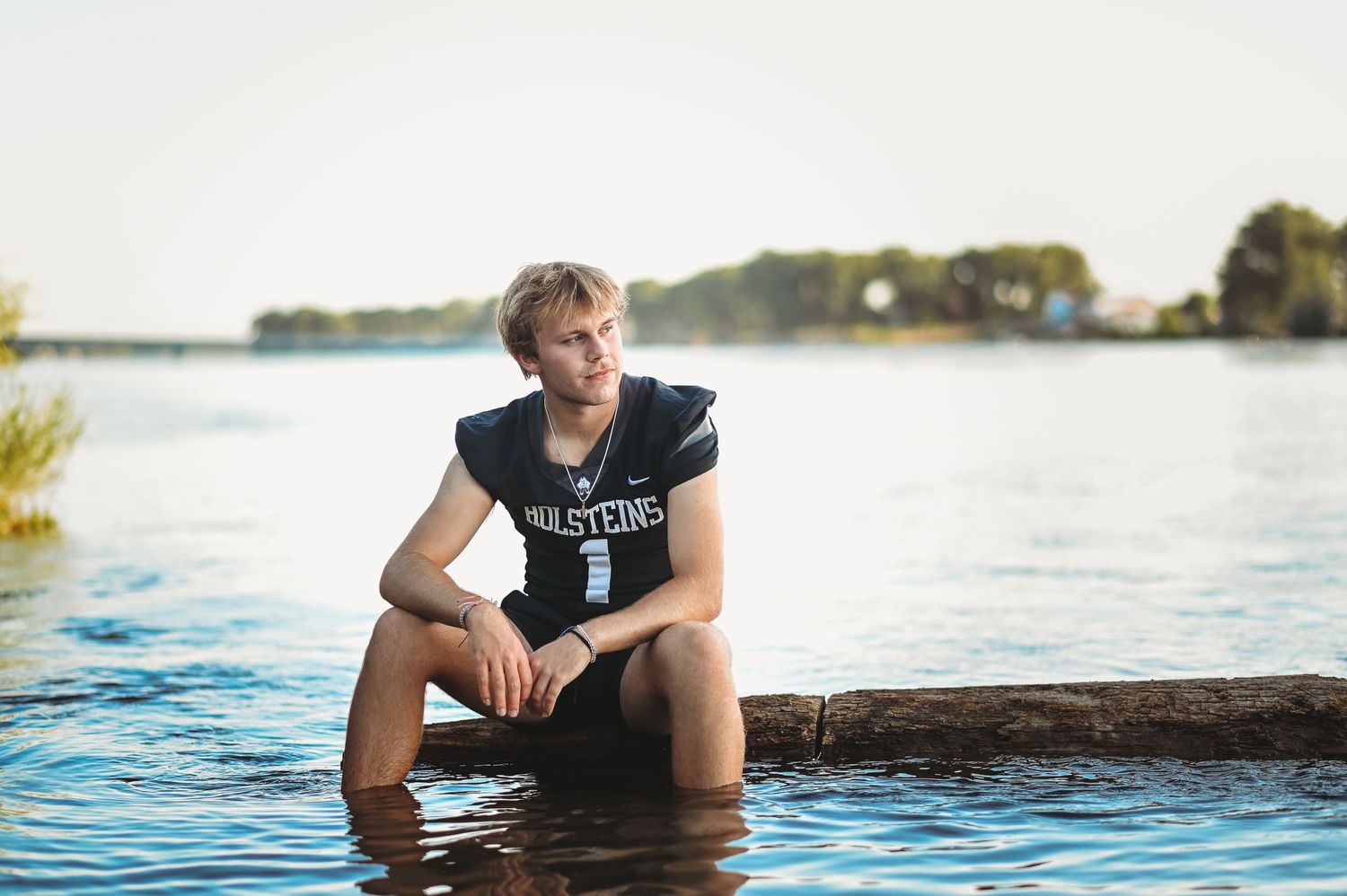 Senior football player sitting on a log at the edge of a peaceful river during sunset.