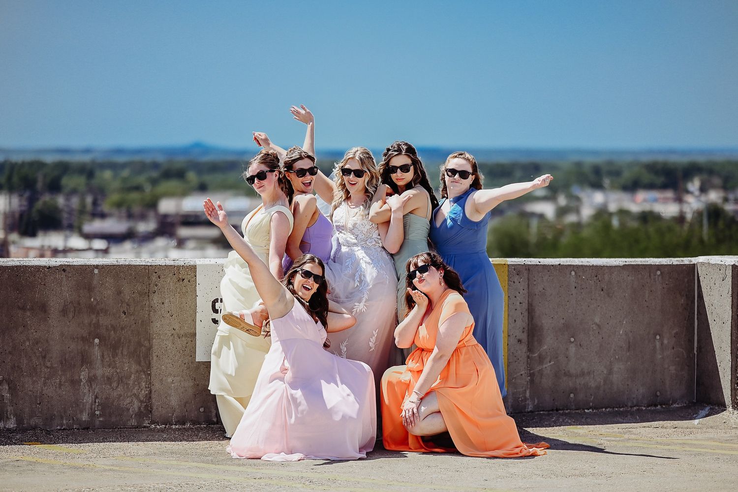 A group in colorful formal attire poses joyfully on a rooftop terrace with a cityscape view in the background.