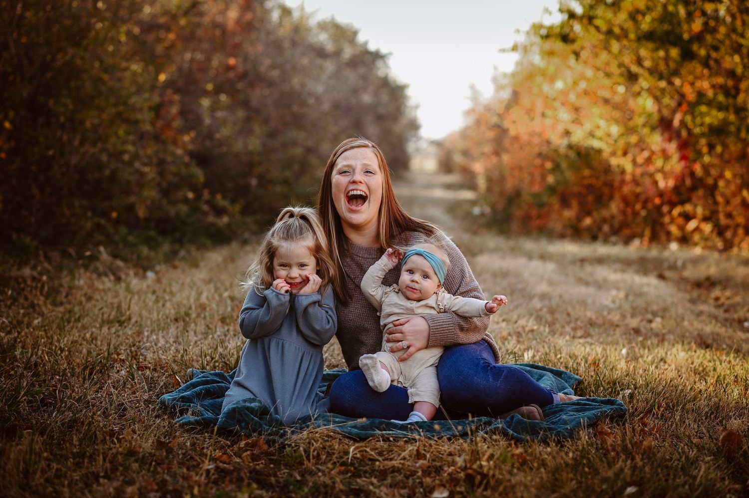 A joyful family moment captured on a blanket during autumn, with colorful fall foliage creating a warm backdrop on a rural path.