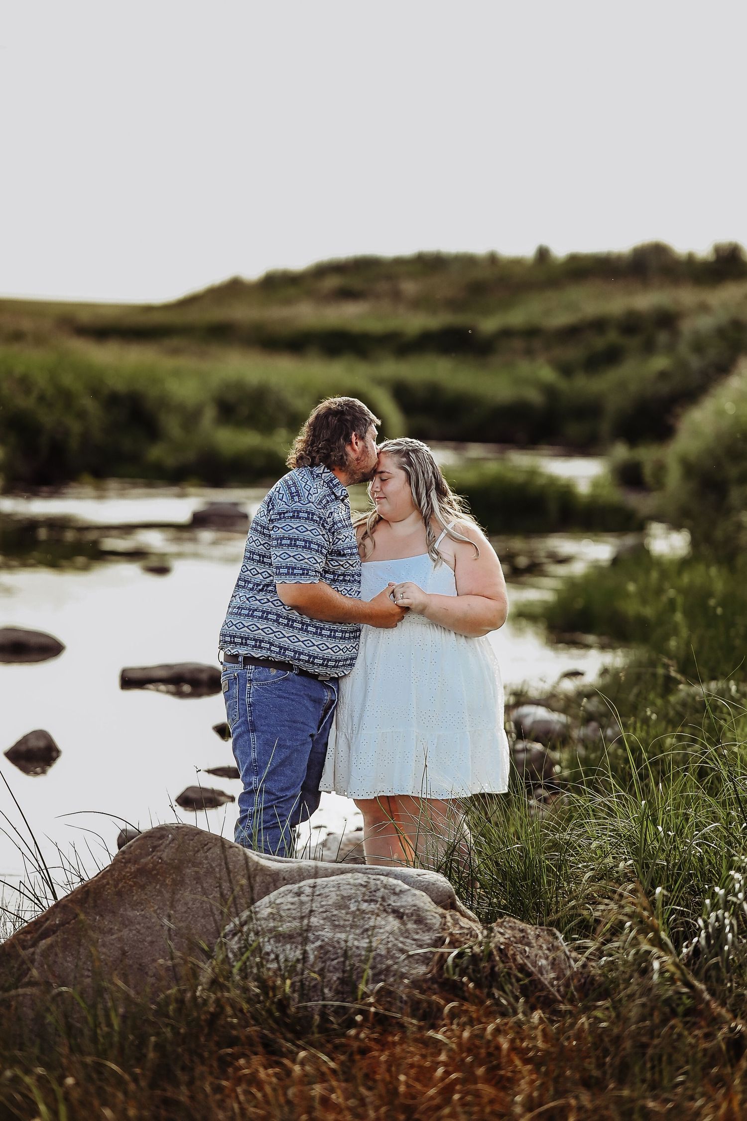 A couple shares an intimate moment by a scenic river with rocks and grass in the foreground during golden hour.