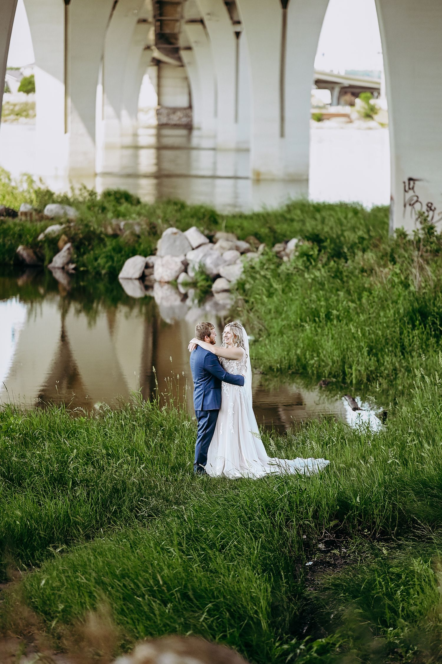 A couple shares a romantic moment under a bridge by the water surrounded by lush green grass and natural scenery.