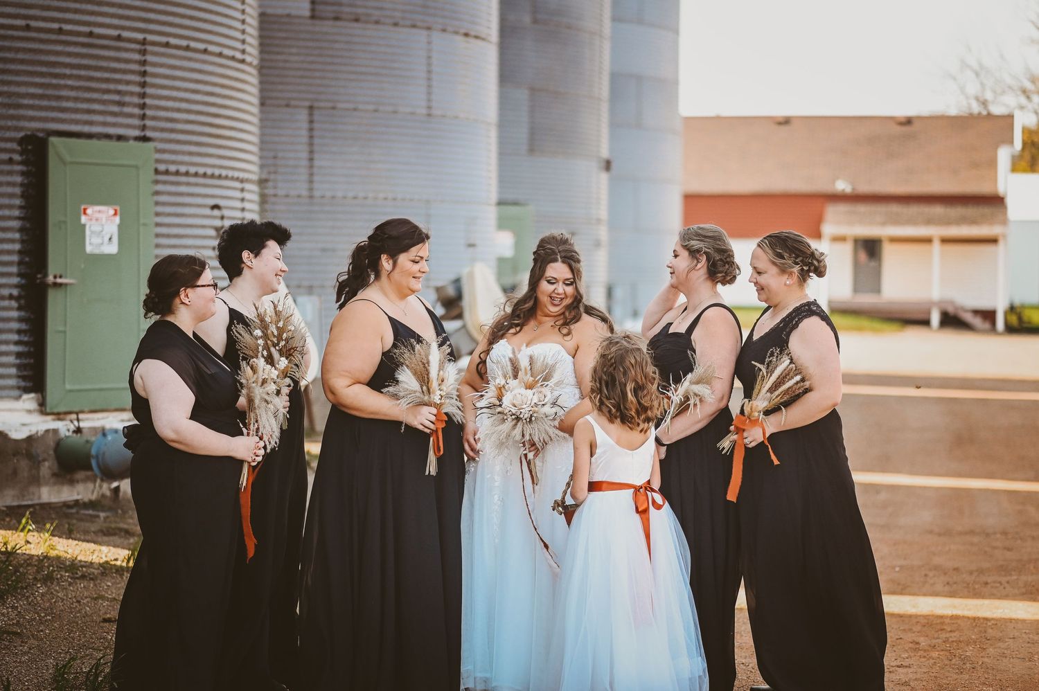 Wedding party in black bridesmaid dresses with orange sashes gather around bride in white gown by rural setting.