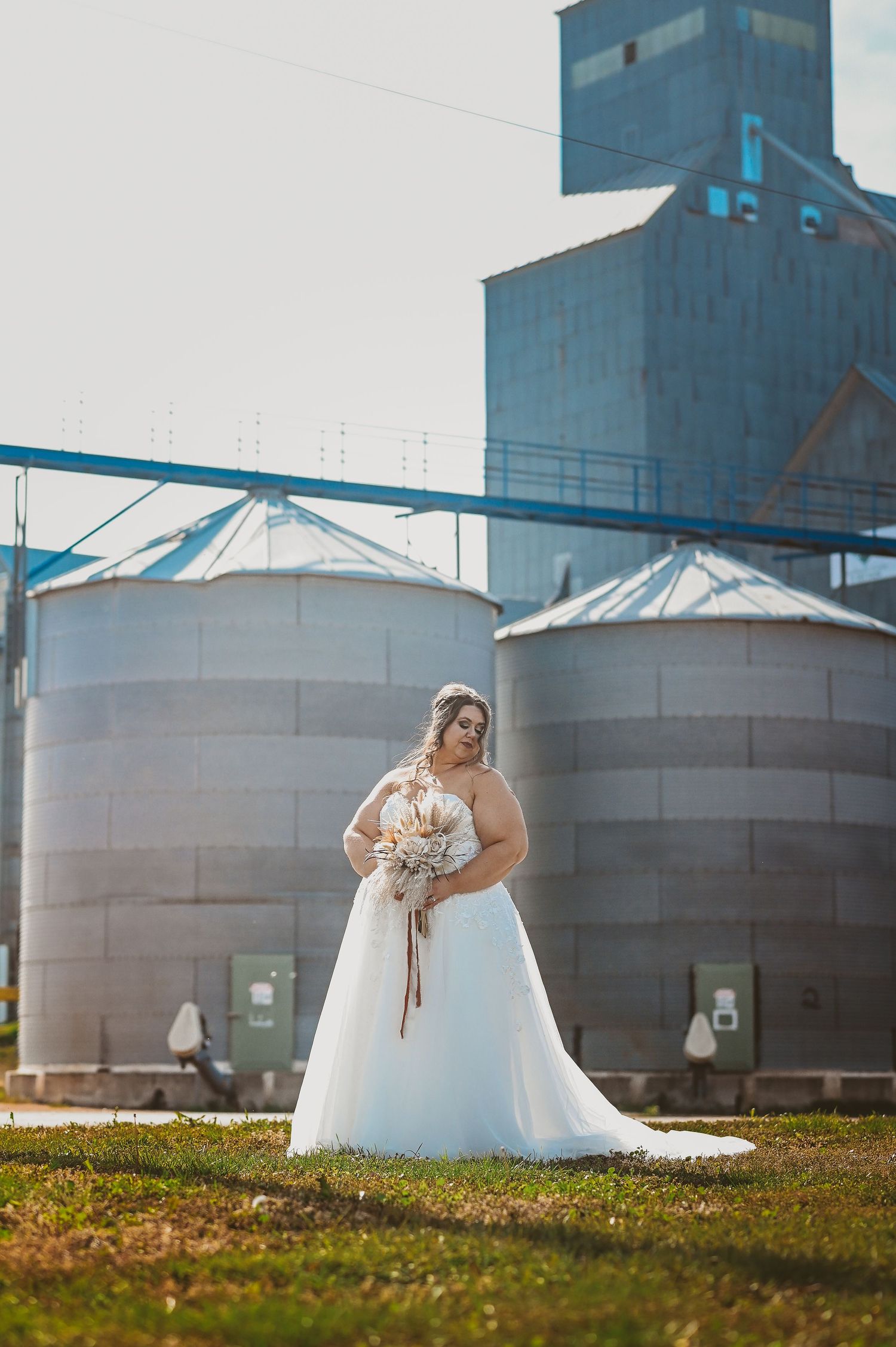 A bride in a flowing white dress poses in front of industrial grain silos and storage tanks on a grassy field.