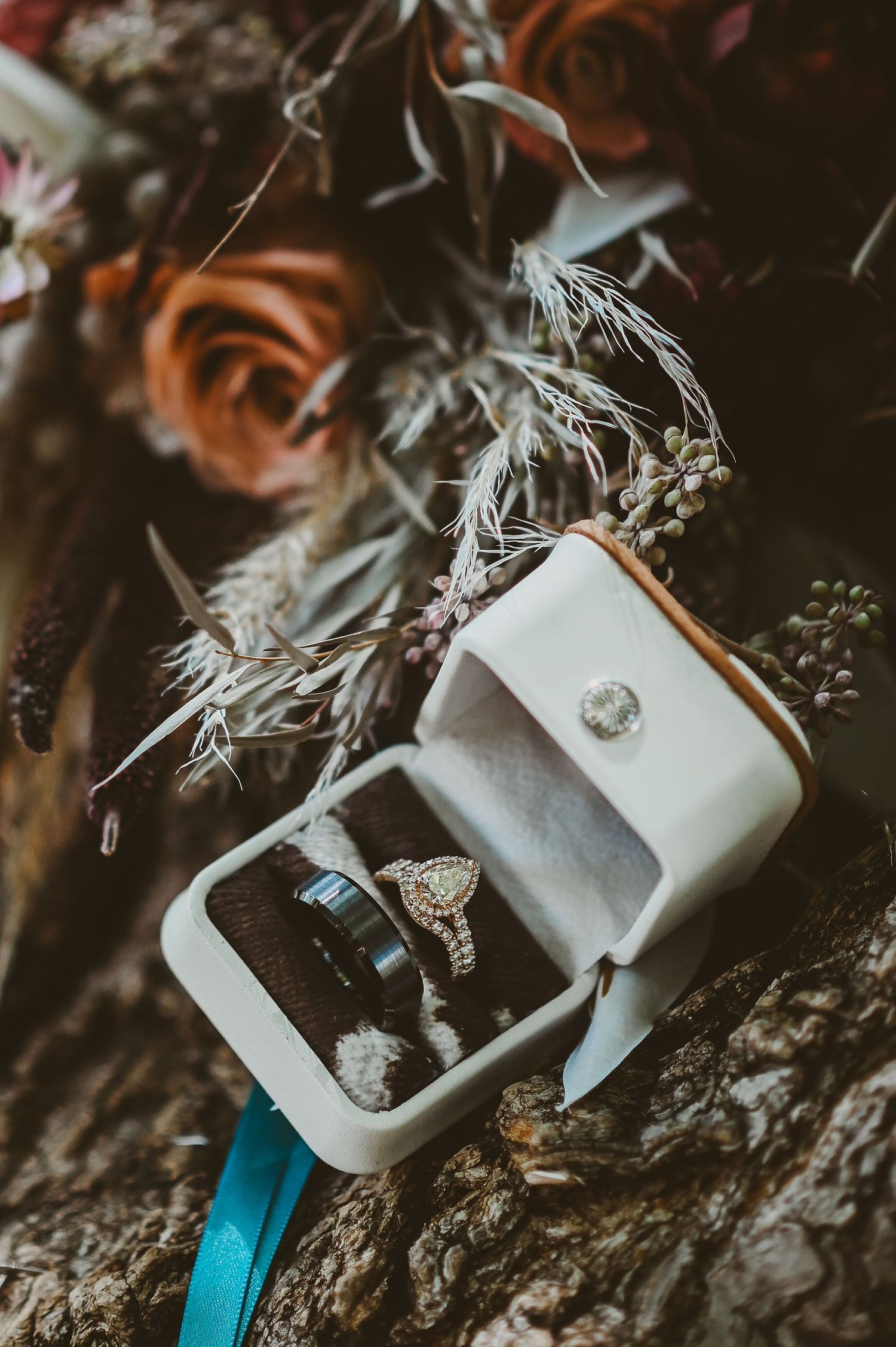 Two diamond rings nestled in a small white ring box surrounded by dried flowers and moody lighting.