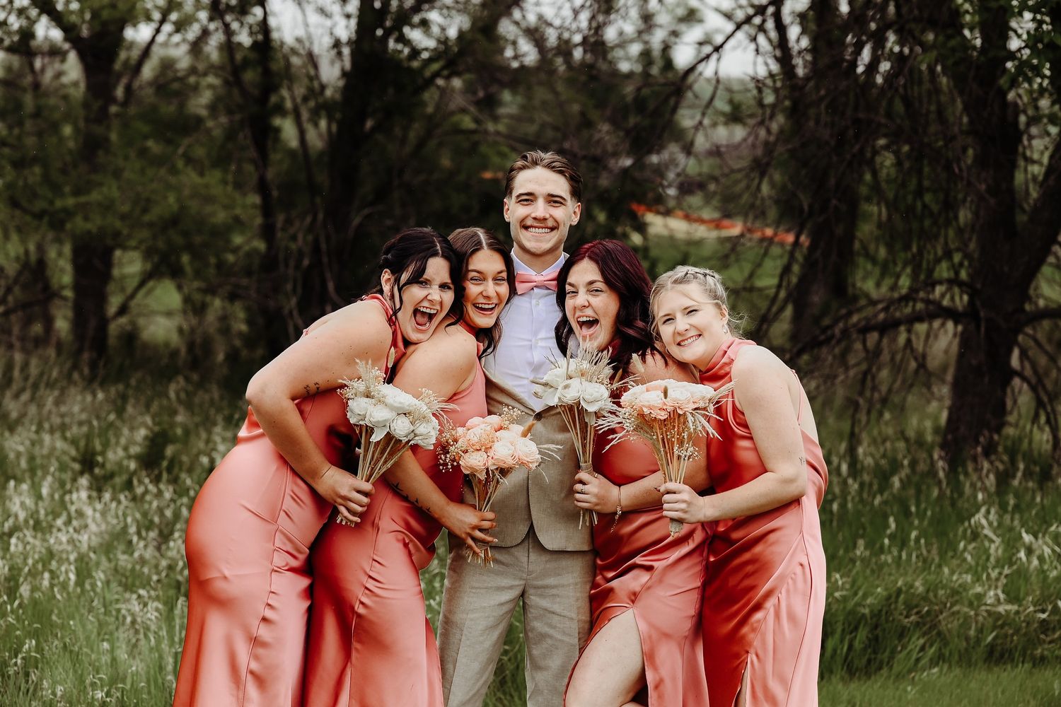 A group of people in coral bridesmaid dresses pose together outdoors in a candid moment.