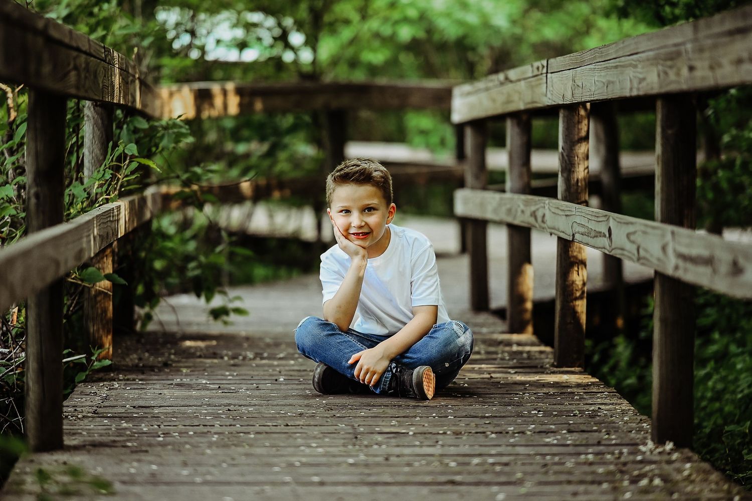 A happy young child in blue sits cross-legged on a wooden pathway surrounded by lush greenery.