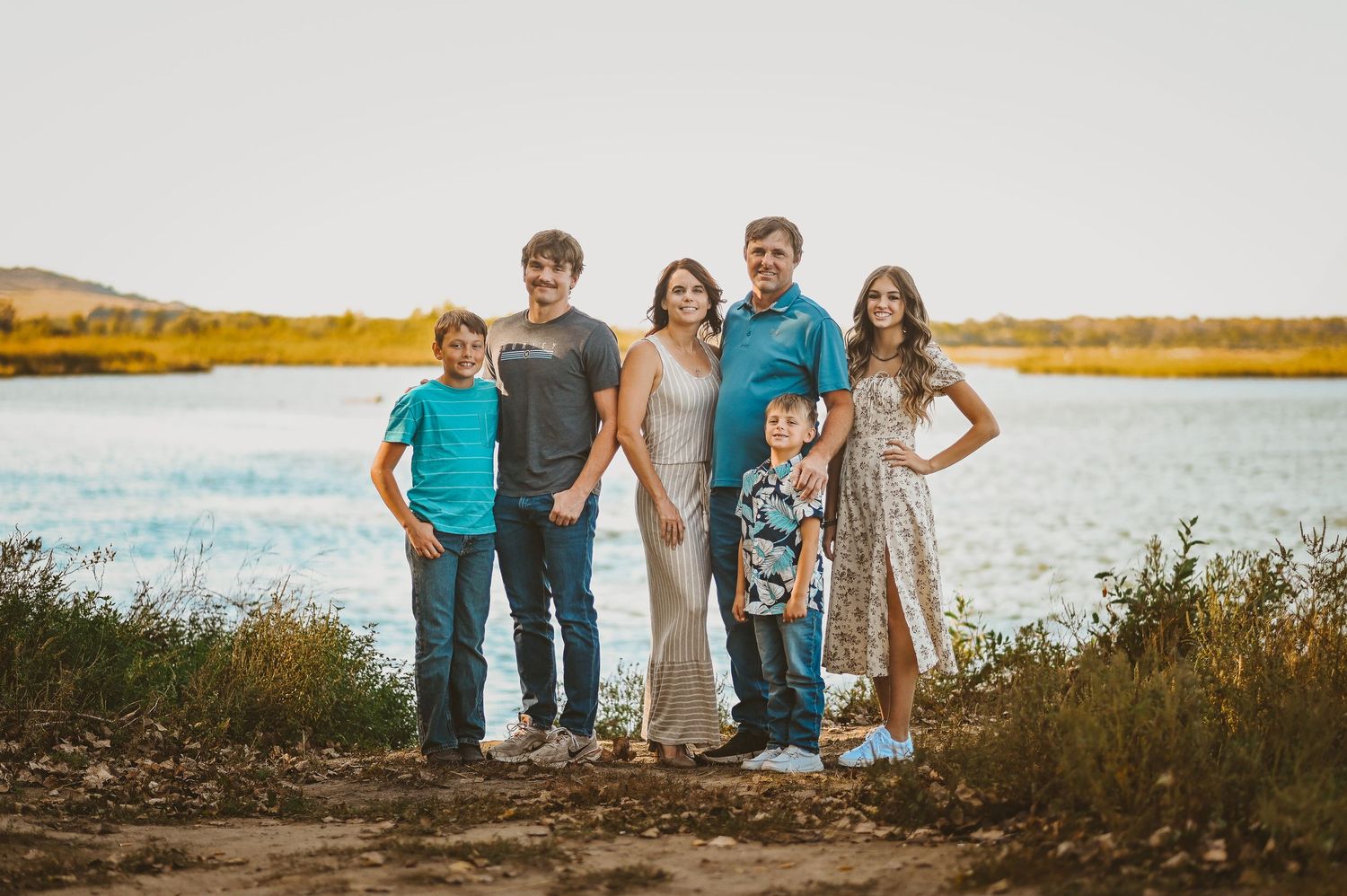A family poses together on the shore of a scenic lake during golden hour with autumn foliage in the background.