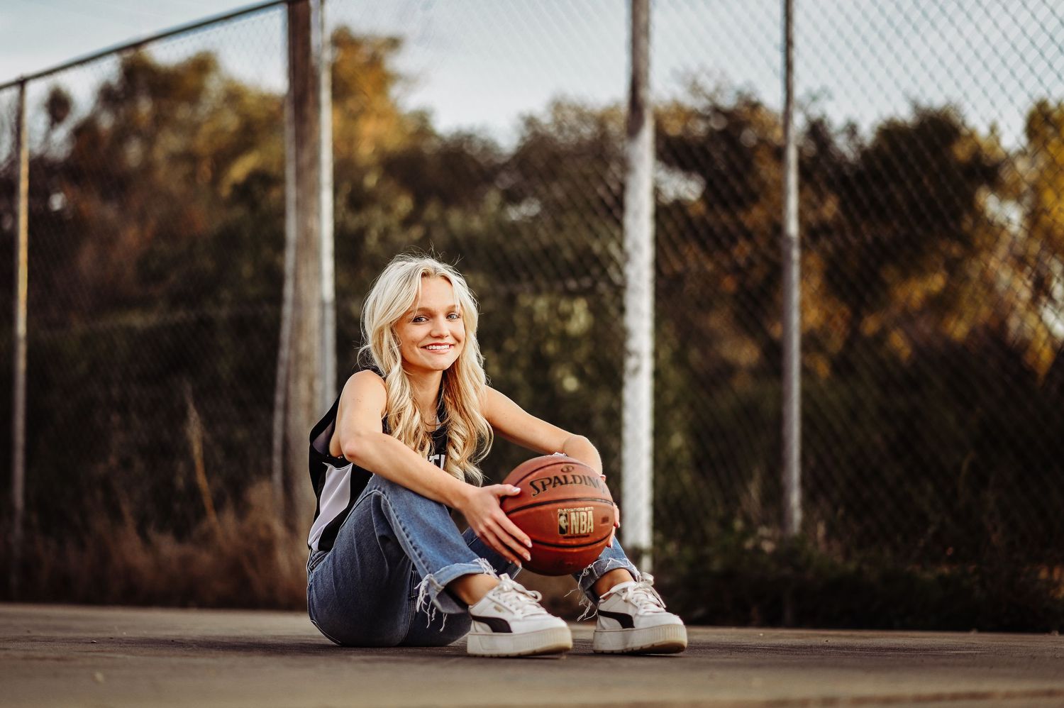 Basketball player in casual attire sits on outdoor court holding a basketball at sunset.