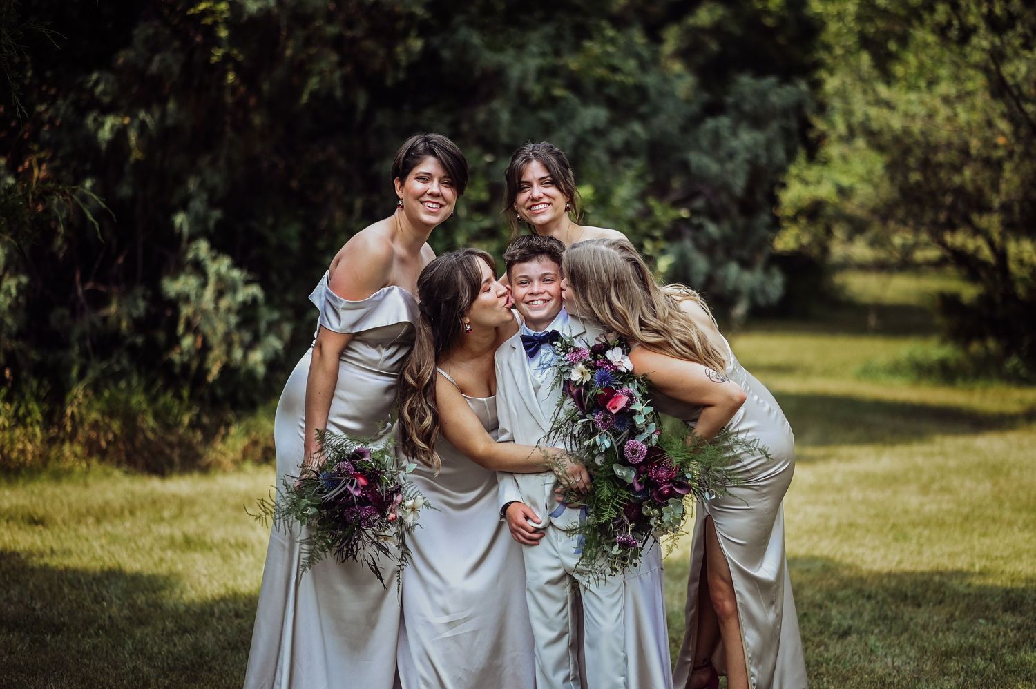 A bridal party in matching rose gold dresses holds purple bouquets while posing and laughing together in a garden setting.