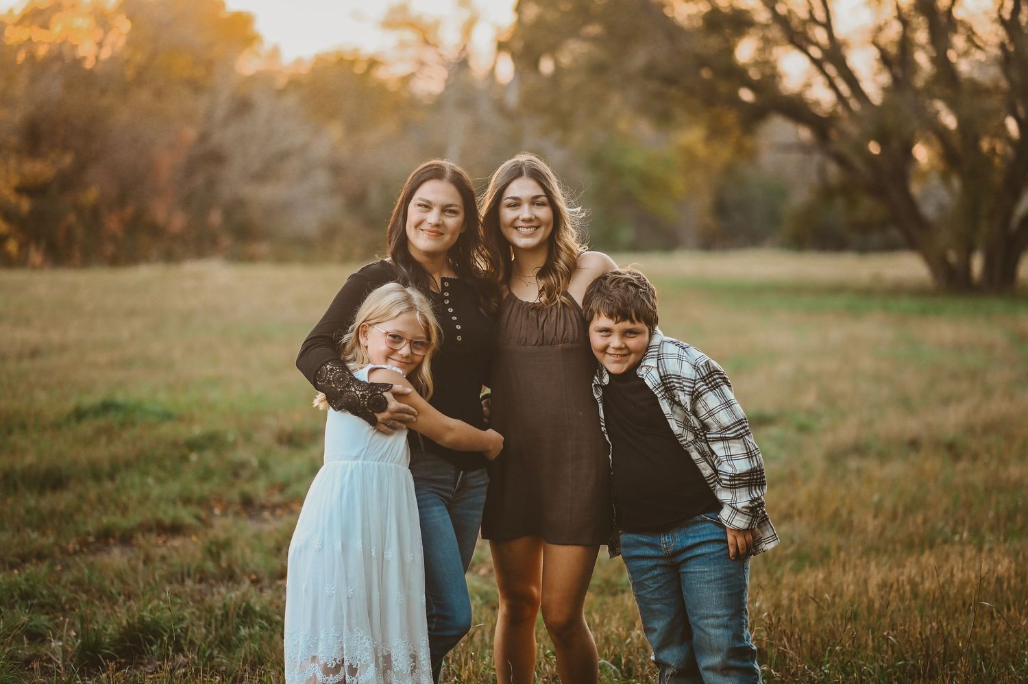 A family group poses together outdoors in a grassy field at golden hour with warm sunlight filtering through trees.