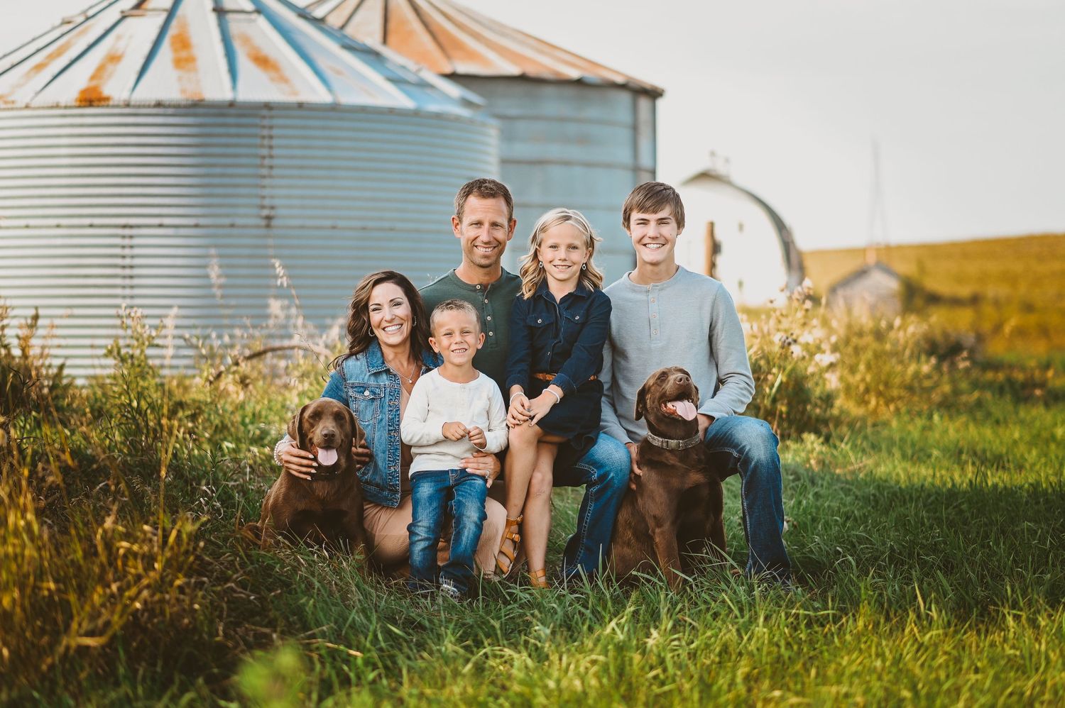 A group poses together with two chocolate Labrador dogs in front of a grain silo at sunset.