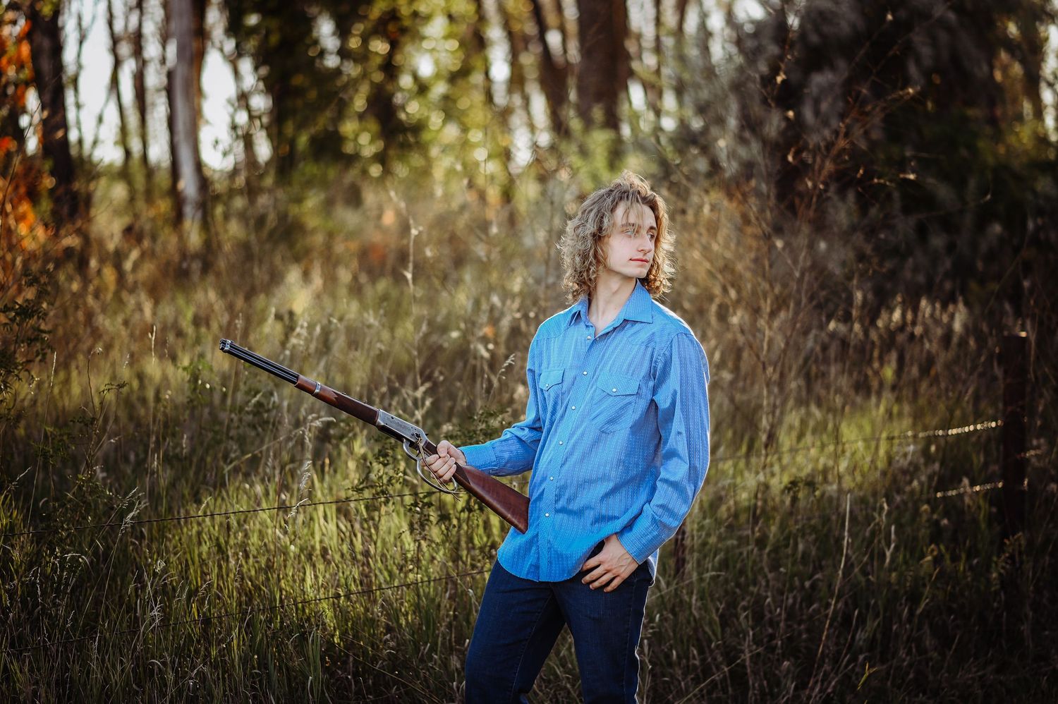 Person in blue button-down shirt holds a rifle while standing in autumn woods during golden hour lighting.