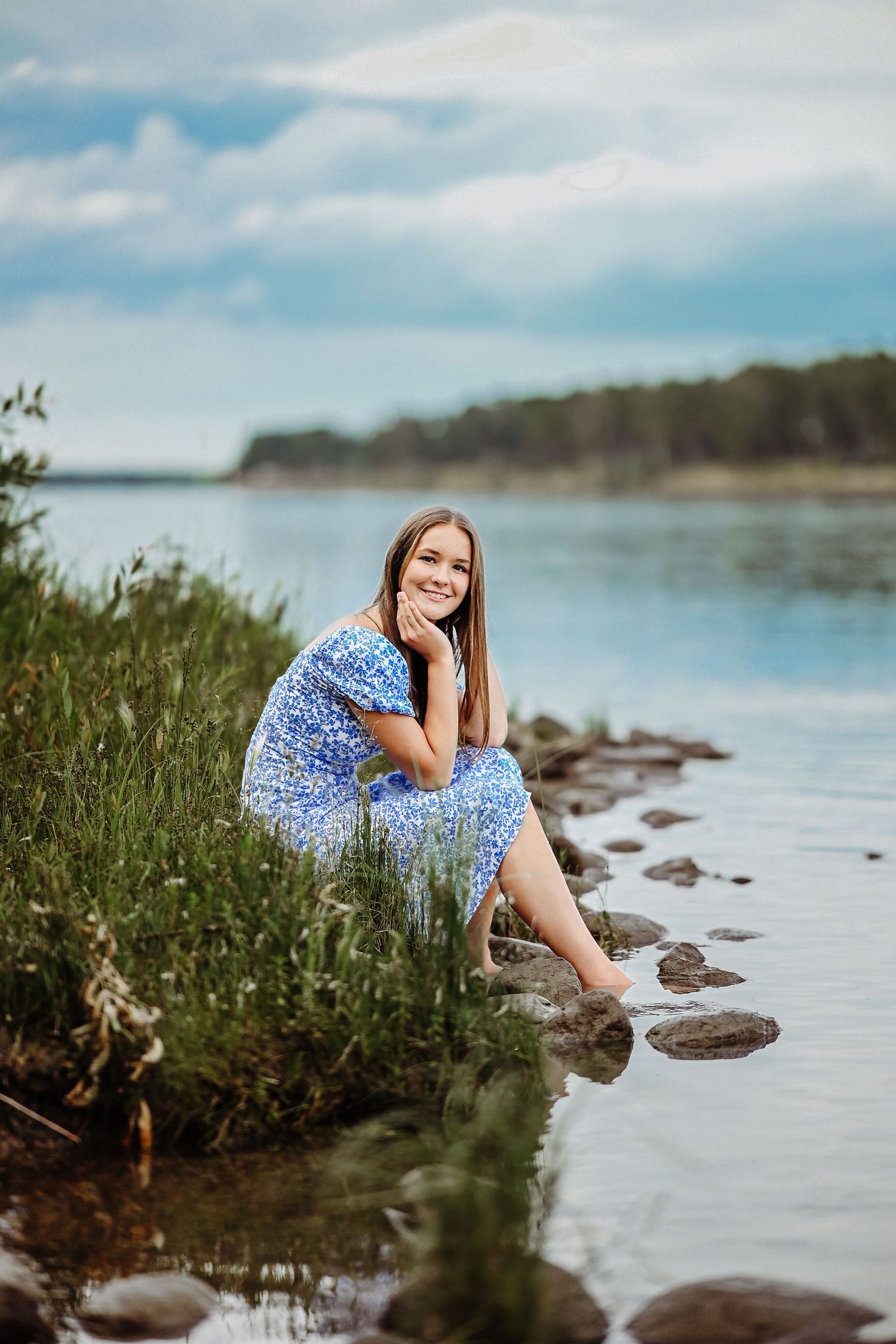 Woman in blue floral dress poses near rocky shoreline with calm water and distant trees on a sunny summer day.