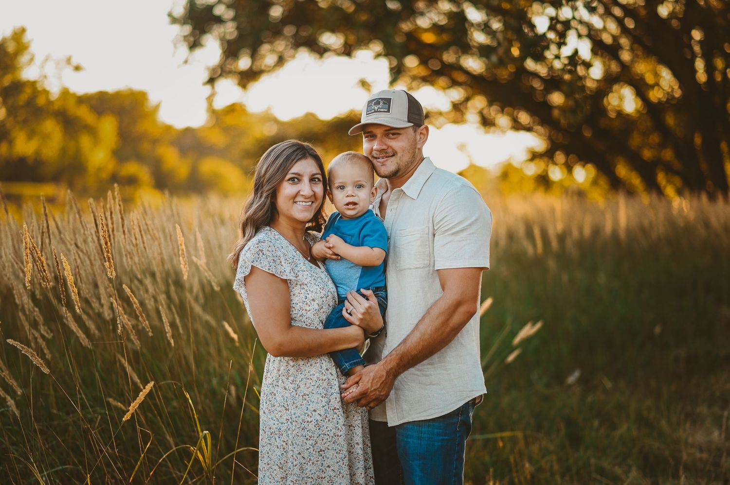 A family poses together in a sunny field at golden hour with tall grass and trees in the background.