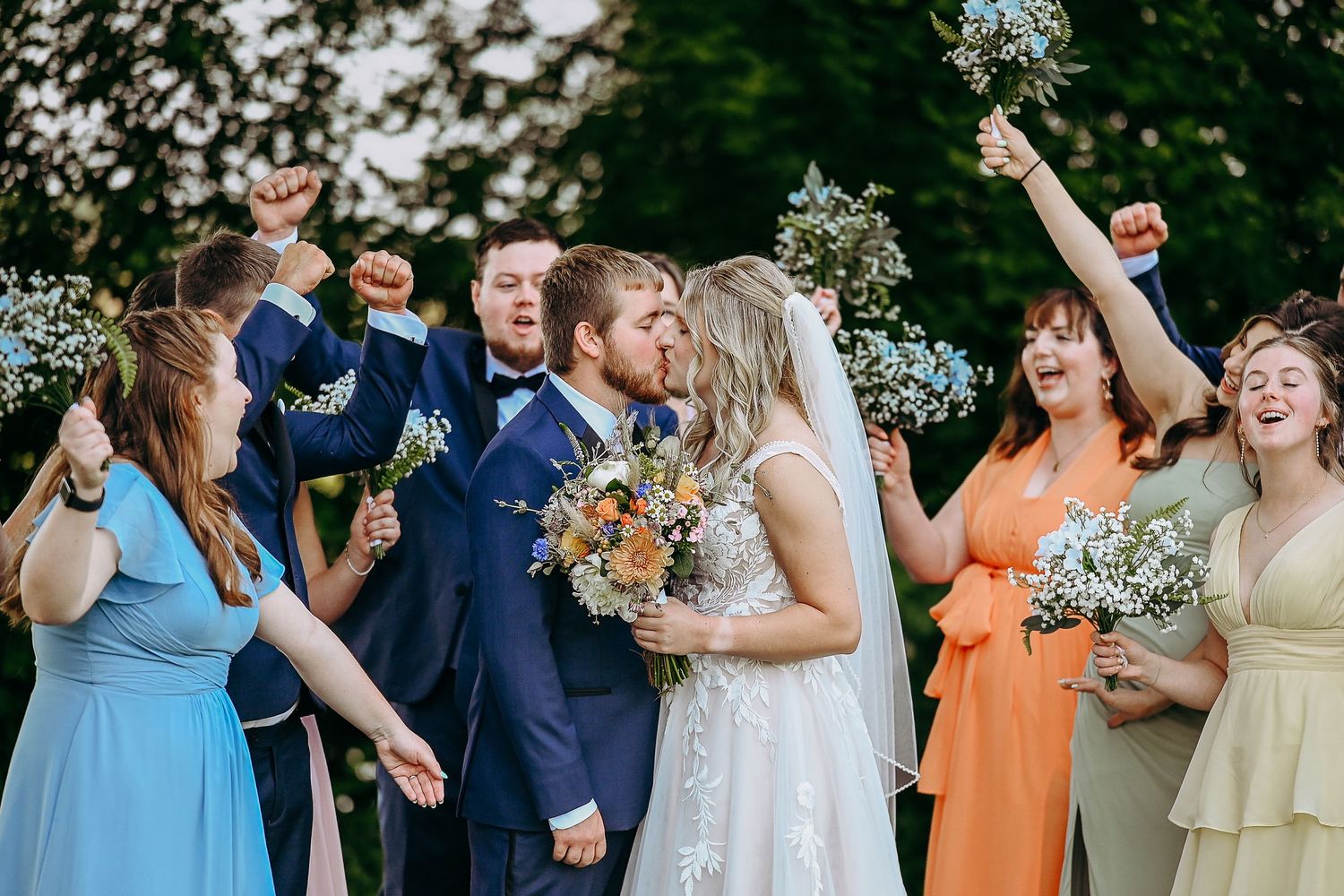 Wedding party celebrates joyfully outdoors surrounded by white floral decorations and wearing colorful attire.
