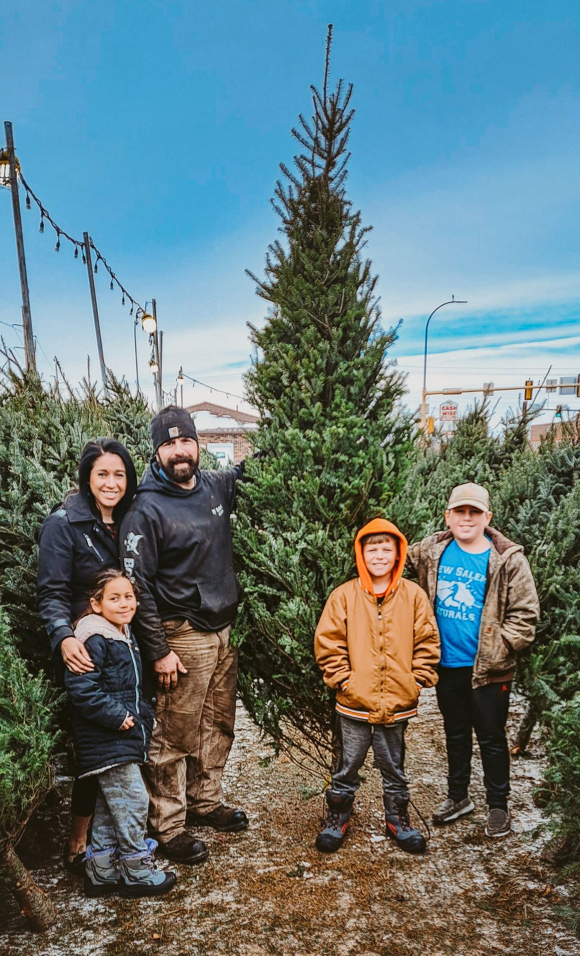 A group stands among evergreen trees at a Christmas tree lot on a bright winter day.