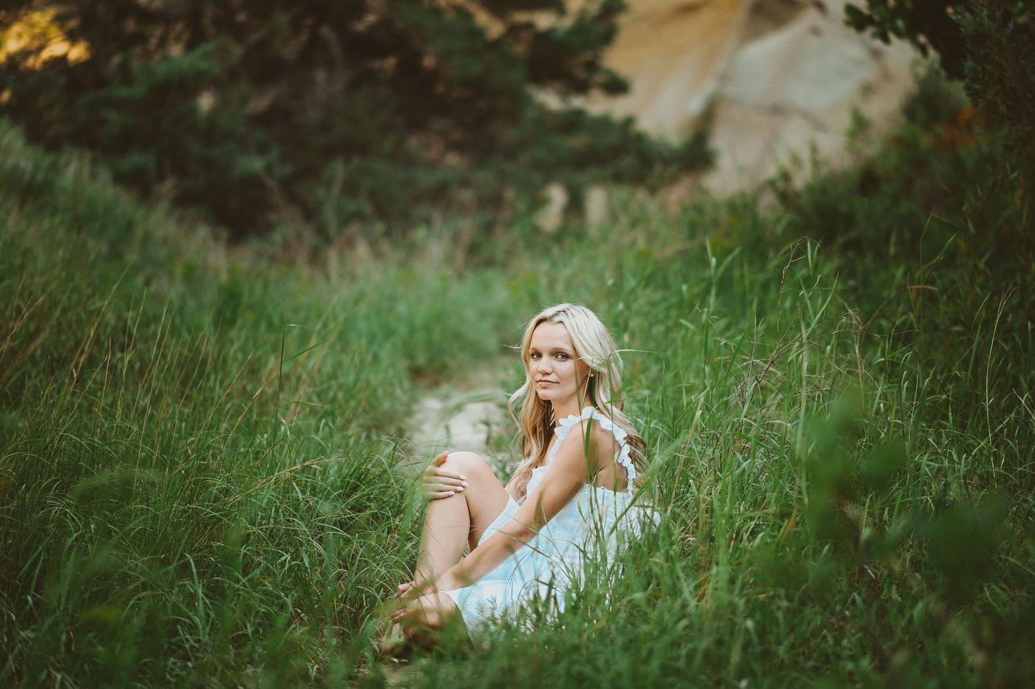 Senior girl in a white dress sits thoughtfully among tall grass in a dreamy outdoor setting during golden hour.